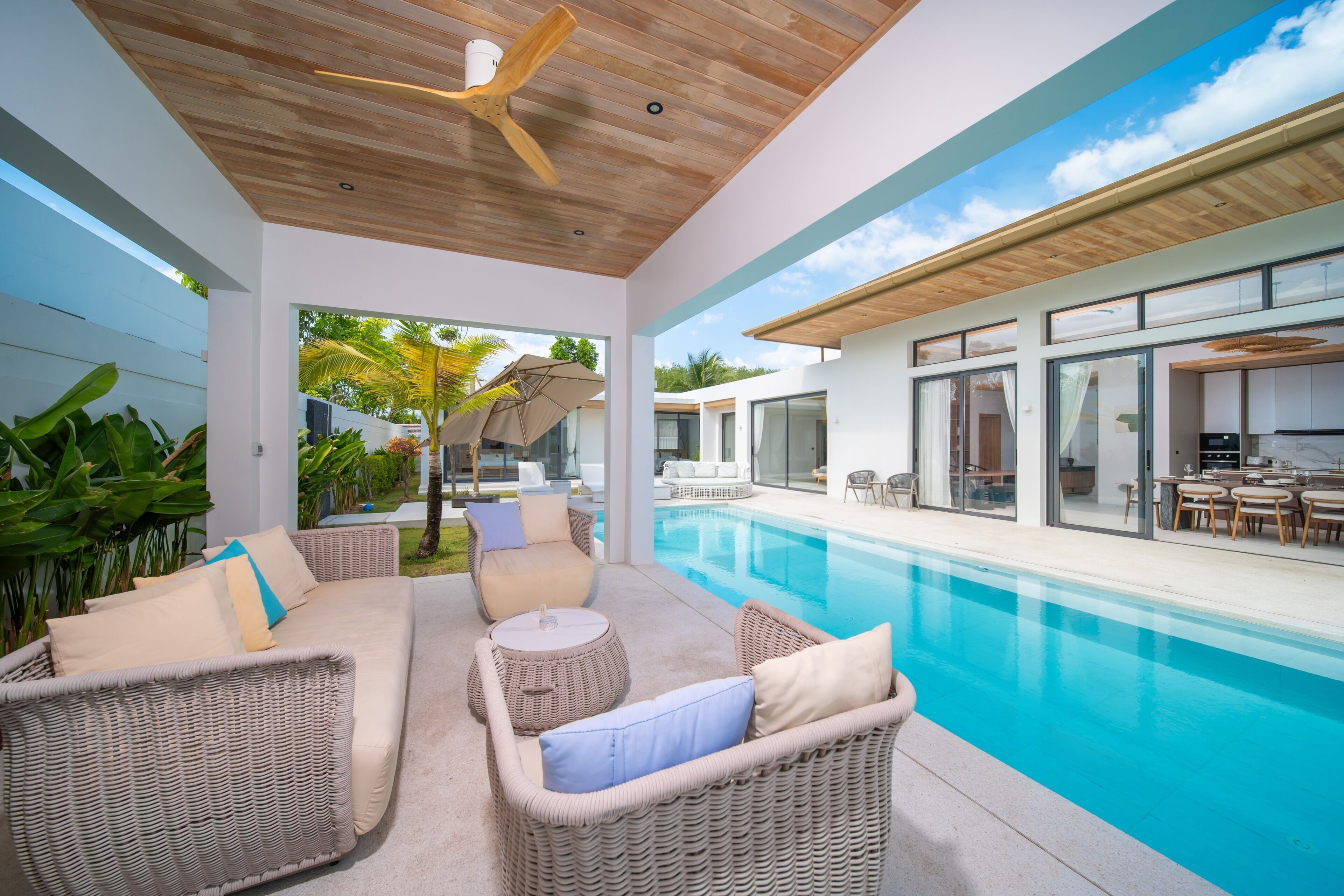 Zona de piscina exterior moderna amb mobiliari de vimet, un ventilador de sostre i vista cap a la casa.