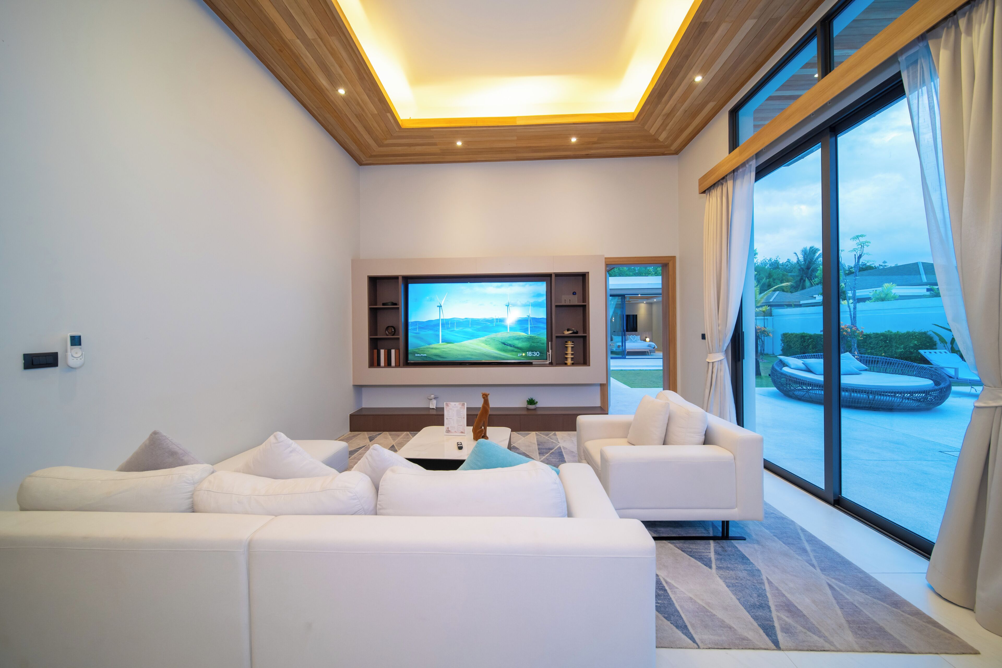 A modern living room with a large flat-screen TV, white sofas, a coffee table, and a view through large windows.