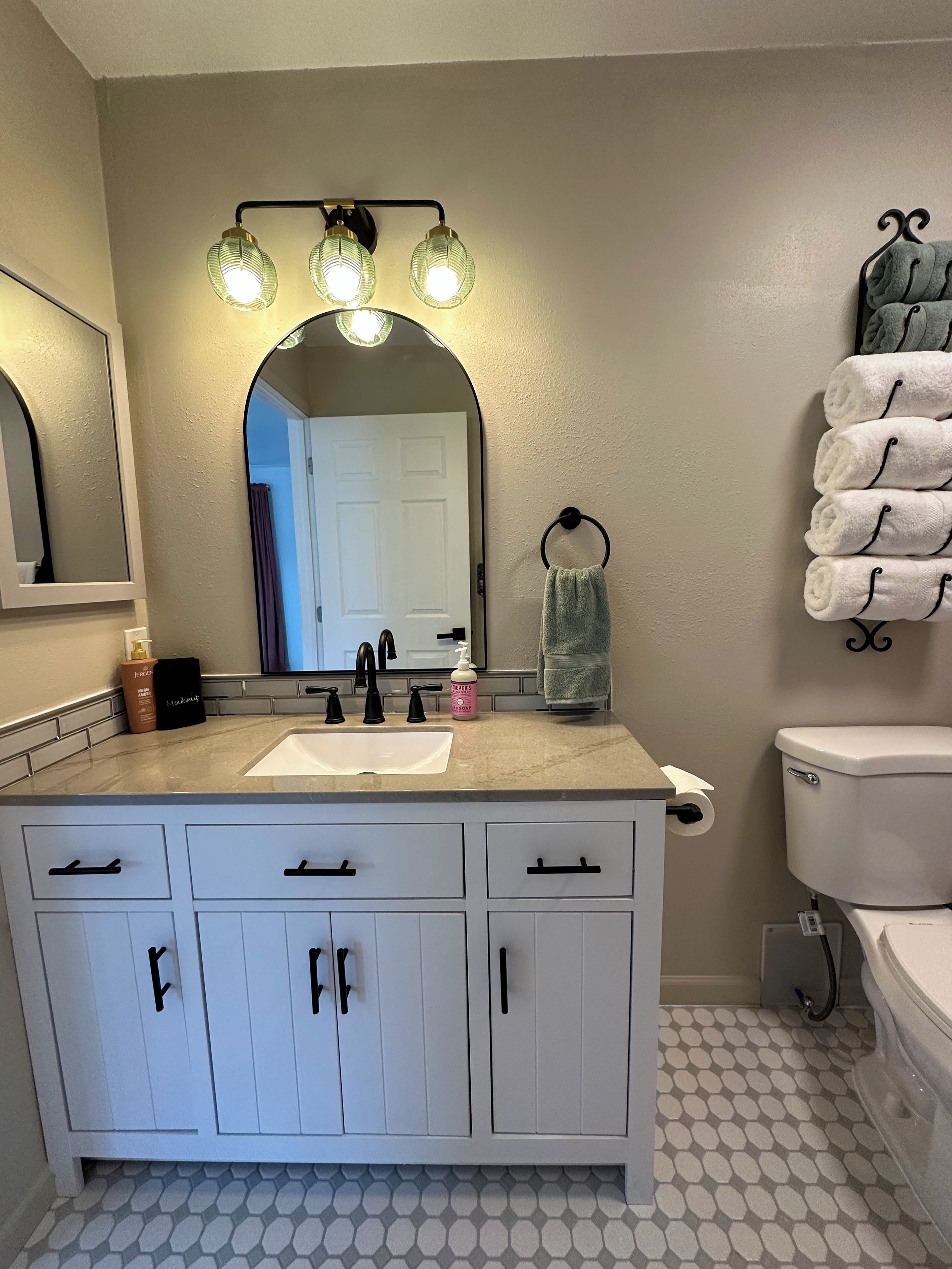 A bathroom with a white vanity, a mirror, a sink, and a toilet. There are towels hanging on a rack and a soap dispenser on the counter.