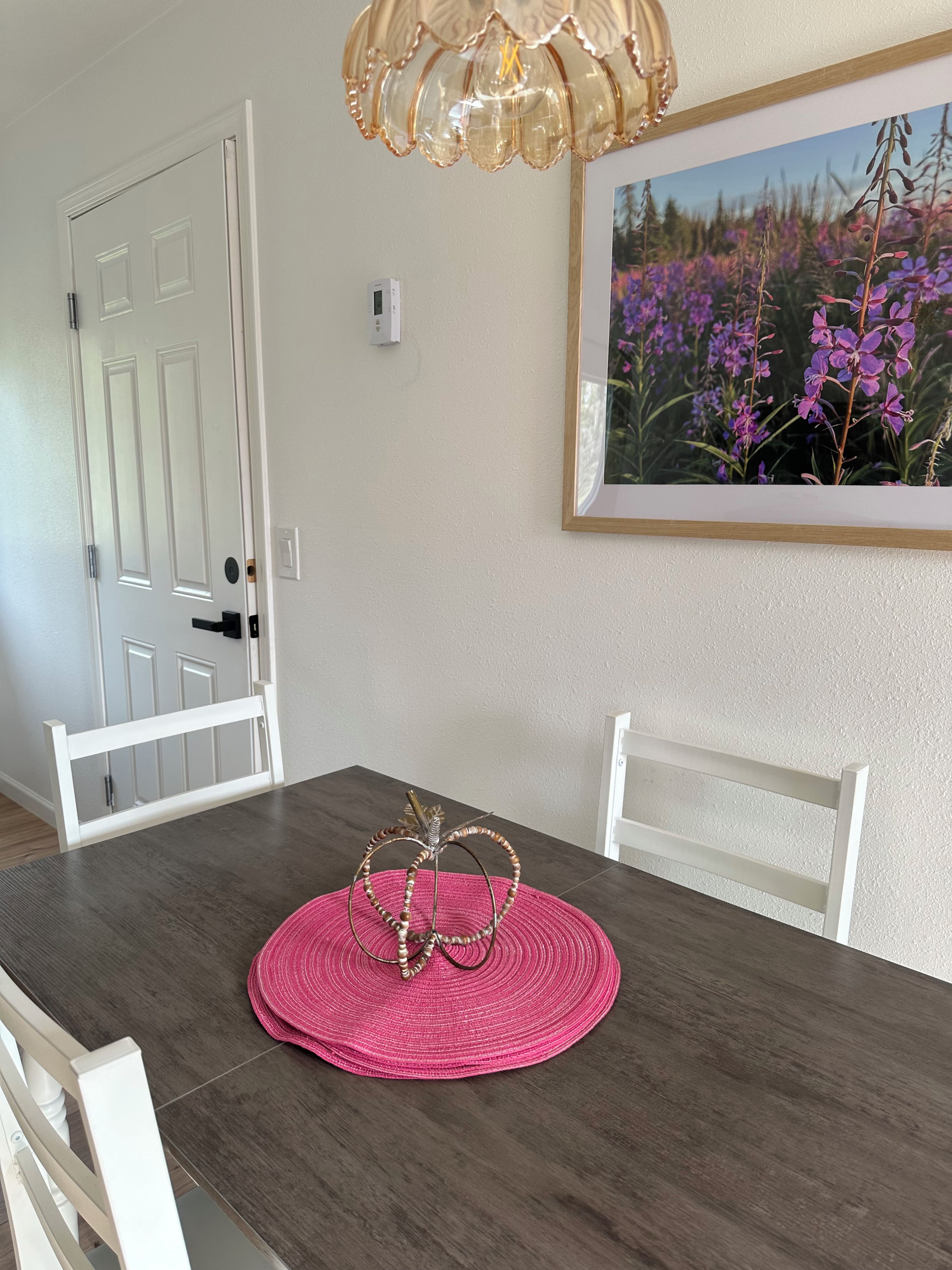 Un comedor con una mesa de madera, sillas blancas, un camino de mesa tejido rosa y un cuadro con flores moradas en la pared.