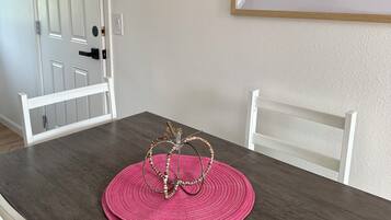 A dining area with a wooden table, white chairs, a pink woven placemat, and a framed picture of purple flowers on the wall.