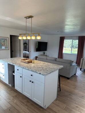 A modern kitchen with a central island, white cabinetry, and a three-light pendant fixture. - Quaint 2-bedroom house in welcoming Kenai for your getaway (Kenai)