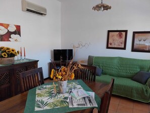 Ein Wohnzimmer mit einem grünen Sofa, einem Holzespeisetisch mit Blumenmuster-Tischdecke, einer Blumenvase und einem Flachbildfernseher.