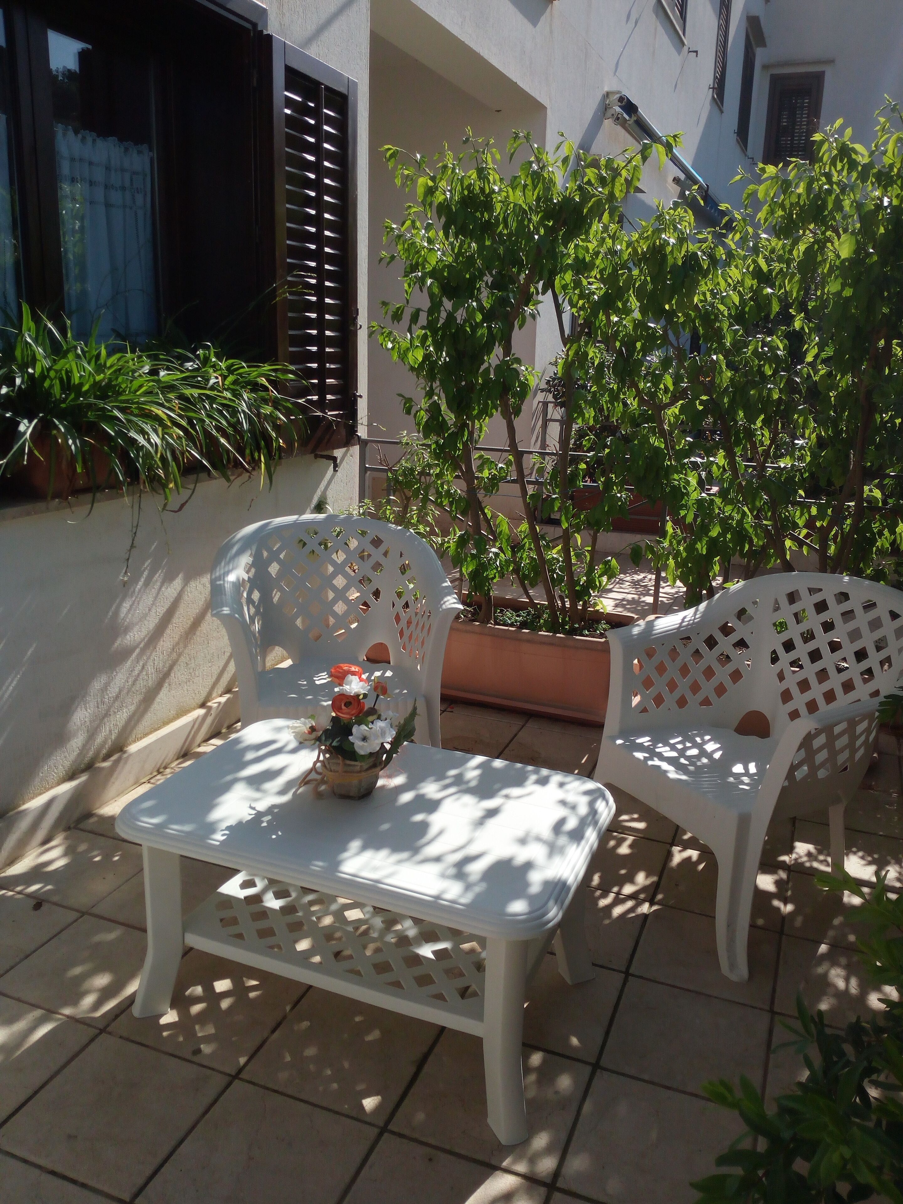 A patio with two white plastic chairs and a small table, surrounded by potted plants and a building in the background.