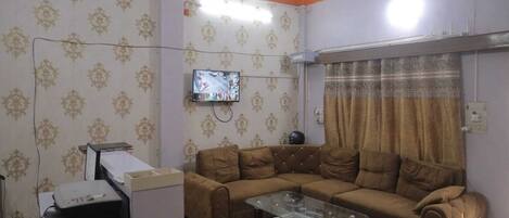 Una sala de estar con un sofá modular, una mesa de centro de vidrio, un televisor montado en la pared y un ventilador de techo.