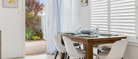 A dining area with a wooden table set for four, white chairs, and a view of a garden through a window with white shutters.