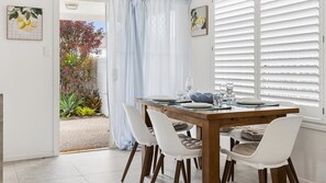 Ein Essbereich mit einem Holztisch, gedeckt für vier Personen, weißen Stühlen und Blick auf einen Garten durch ein Fenster mit weißen Fensterläden.