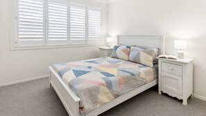 A bedroom with a bed, two bedside tables, a nightstand, a window with blinds, and a carpeted floor. - Baillie s Getaway - Lifestyle Yamba (YAMBA)
