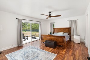 Ein Schlafzimmer mit einem hölzernen Bett, einem Deckenventilator, einem großen Fenster mit Vorhängen und Blick auf Grünfläche.
