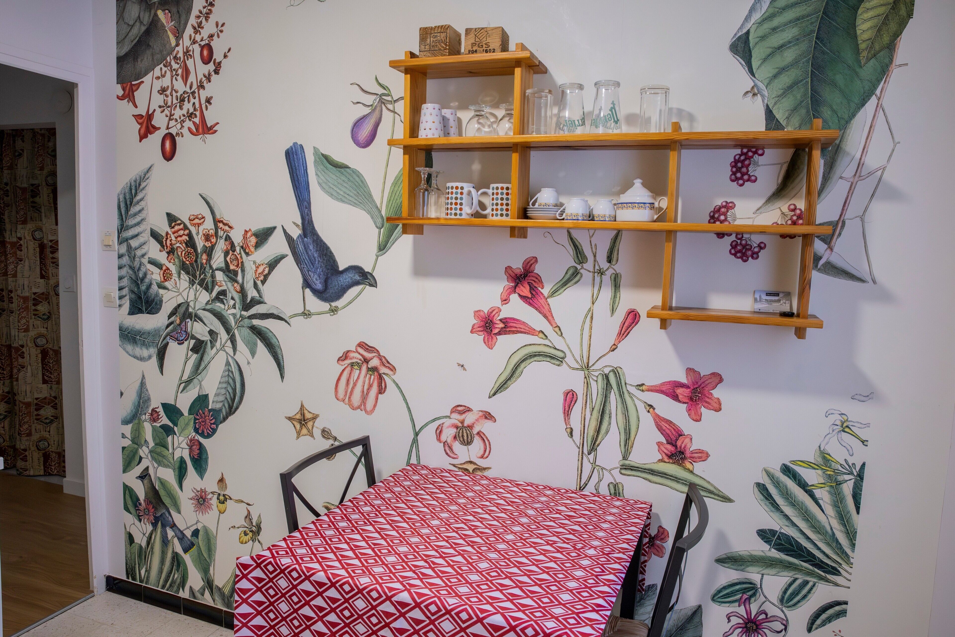 Ein Zimmer mit einer Wandmalerei von tropischen Pflanzen, einem Holzregal mit Gläsern und einer rot gemusterten Tischdecke.