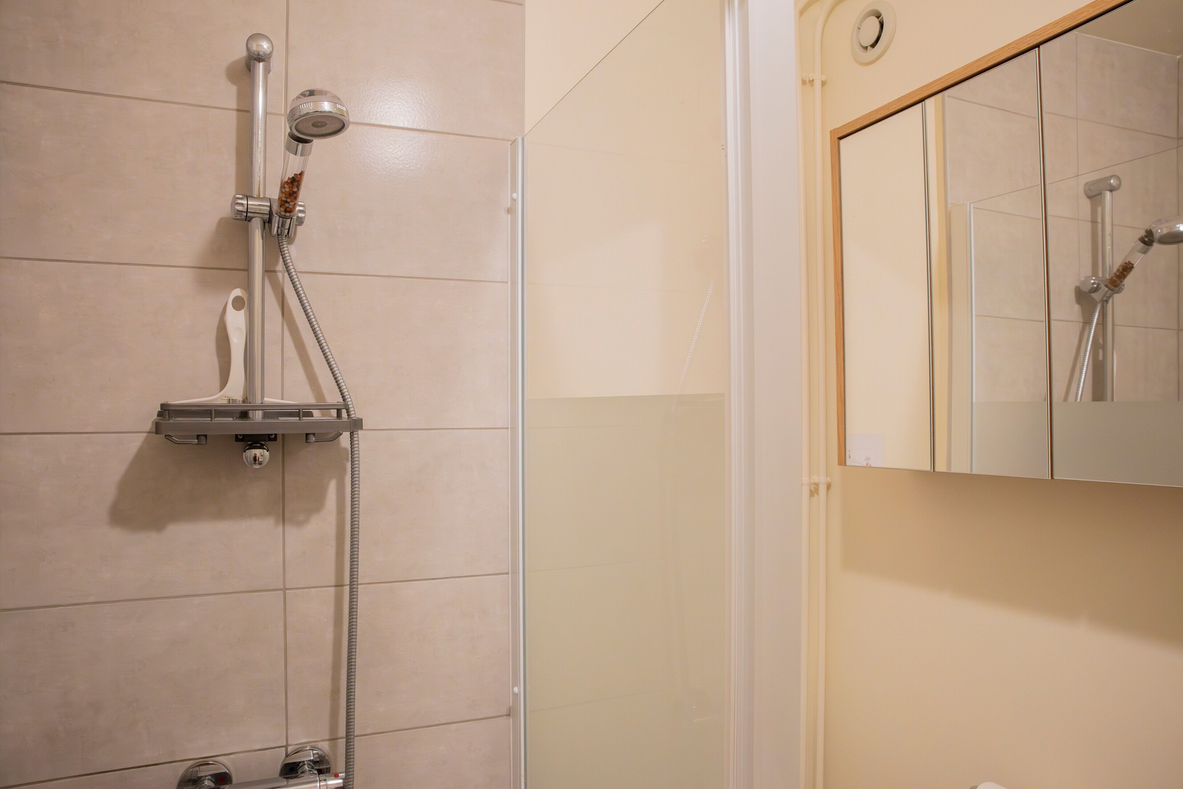 A shower with a handheld showerhead and a wall-mounted faucet, a mirror above the sink, and a tiled wall.