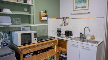 A compact kitchen with a microwave, coffee maker, and sink. There are wooden crates with dishes and a small trash bin.