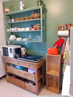 A kitchen with a microwave, kettle, and coffee maker on a wooden counter. There is a green shelf with various kitchen items and a basket. A small wooden stool with a red object on top is next to the counter. - Accommodation for 2 with Pool (Cour-Cheverny)