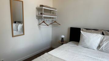 A bedroom with a bed, a hanging shelf with towels, a mirror, and a bedside table with a lamp.