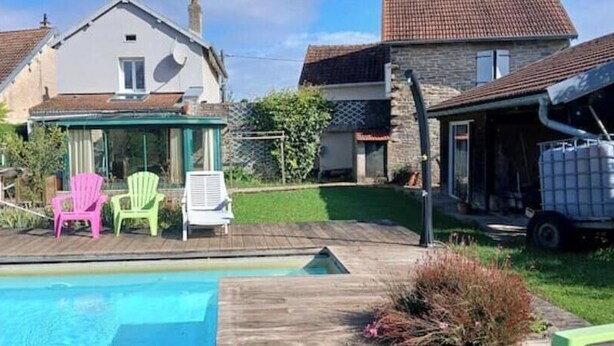 A swimming pool with a wooden deck, two green and one pink chair, a white chair, and a truck parked in the background. - Gîte Familial au Coeur du Triangle Vert - Piscine et Détente Assurées (Borey)