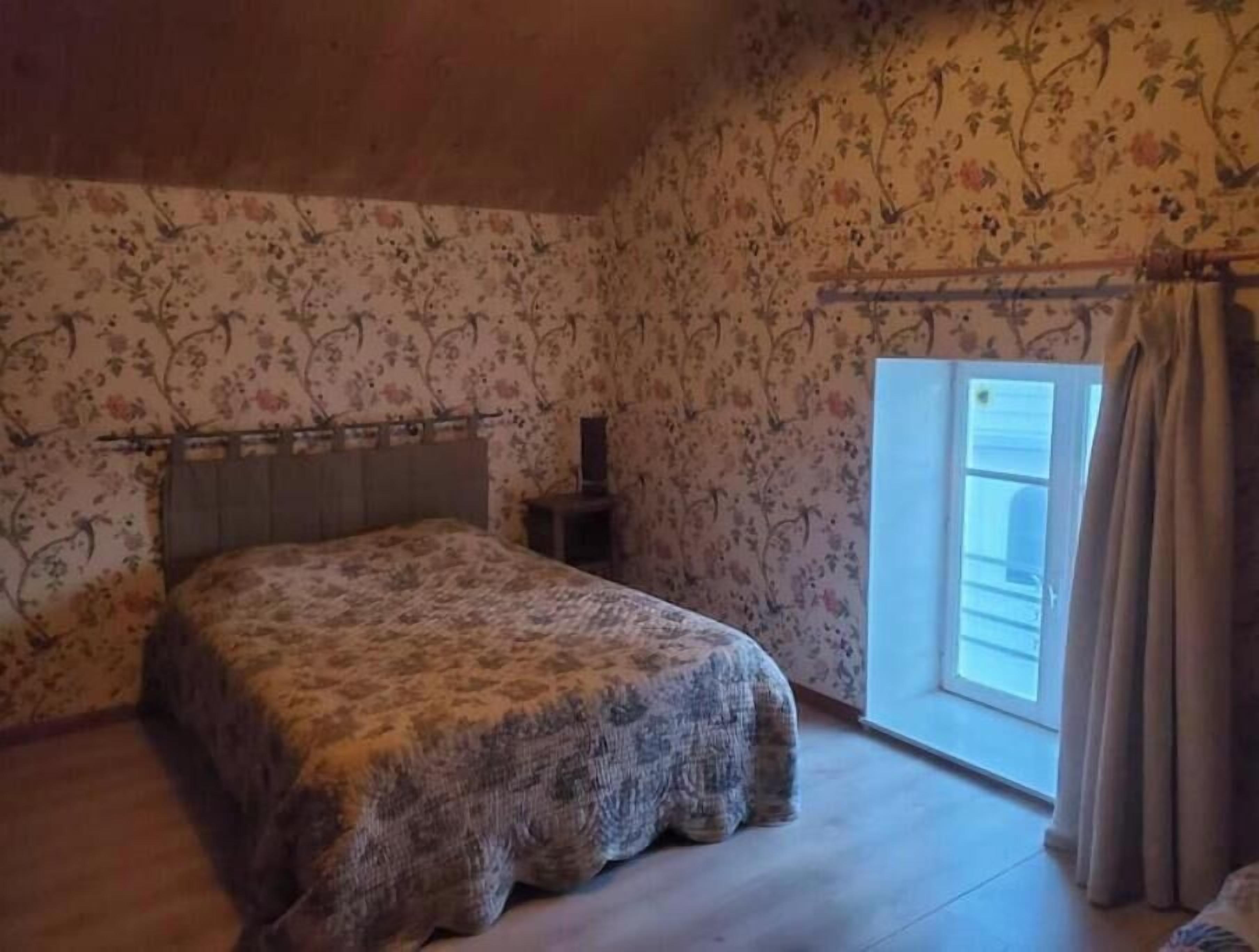 A bedroom with a bed, a nightstand, a window with curtains, and floral wallpaper.