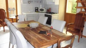 A dining area with a wooden table and white chairs, a kitchen with white cabinets, and a view of greenery outside. - Gîte Familial au Coeur du Triangle Vert - Piscine et Détente Assurées (Borey)