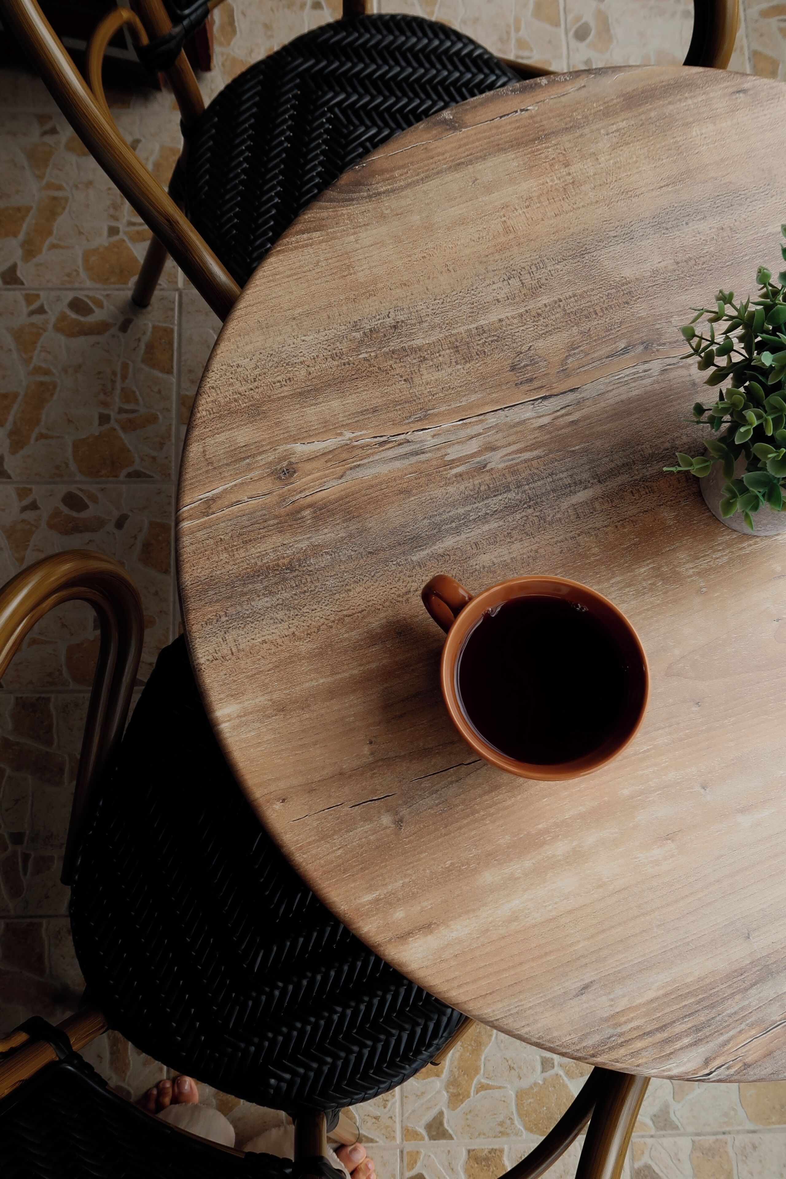 Une table ronde en bois, sur laquelle se trouvent une tasse de café, une petite plante en pot et une chaise en osier.