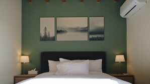 A neatly made bed with white linens, two bedside lamps, and three framed landscape paintings on the wall. - Cozy mountain apartment in Platres (Pano Platres)
