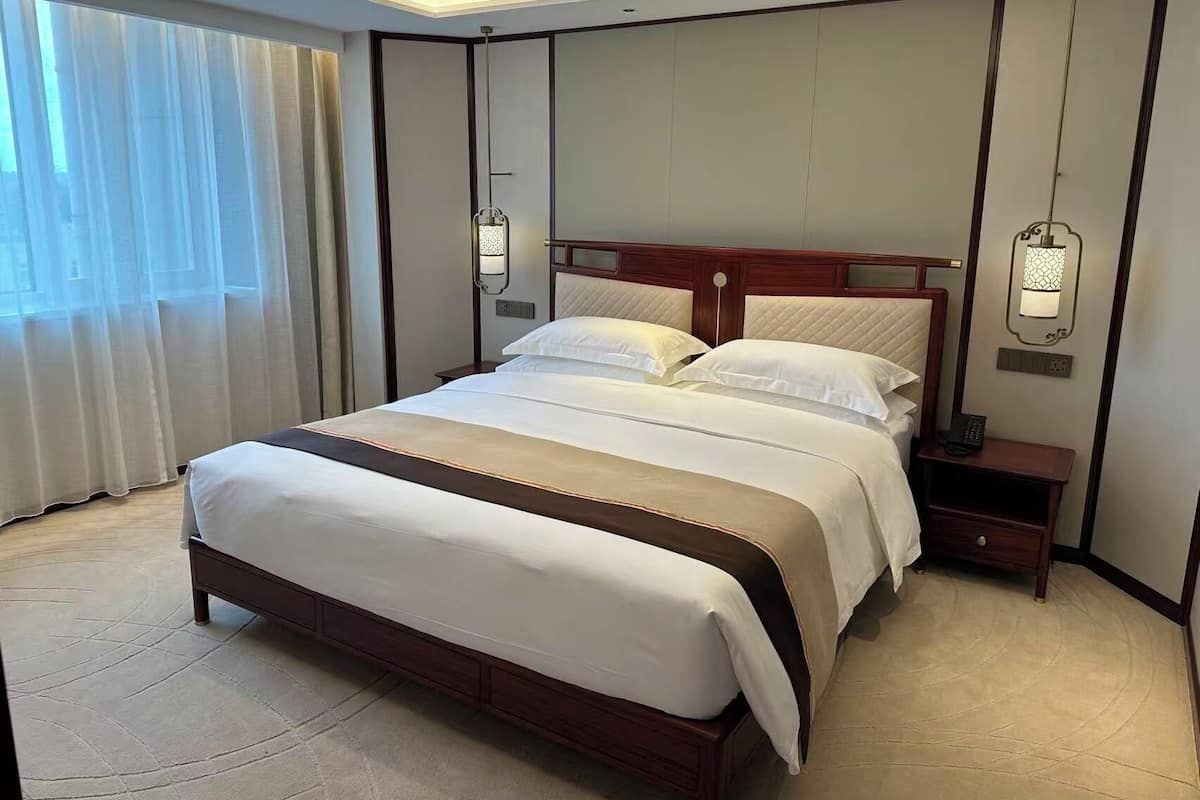 A hotel room with a large bed, bedside tables, a hanging lamp, and a window with sheer curtains.