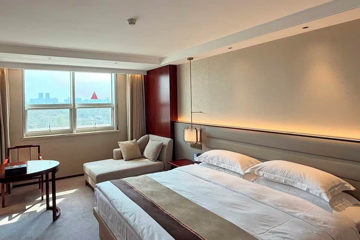 A hotel room with a large bed, a desk, a chair, and a view of a cityscape through the window.