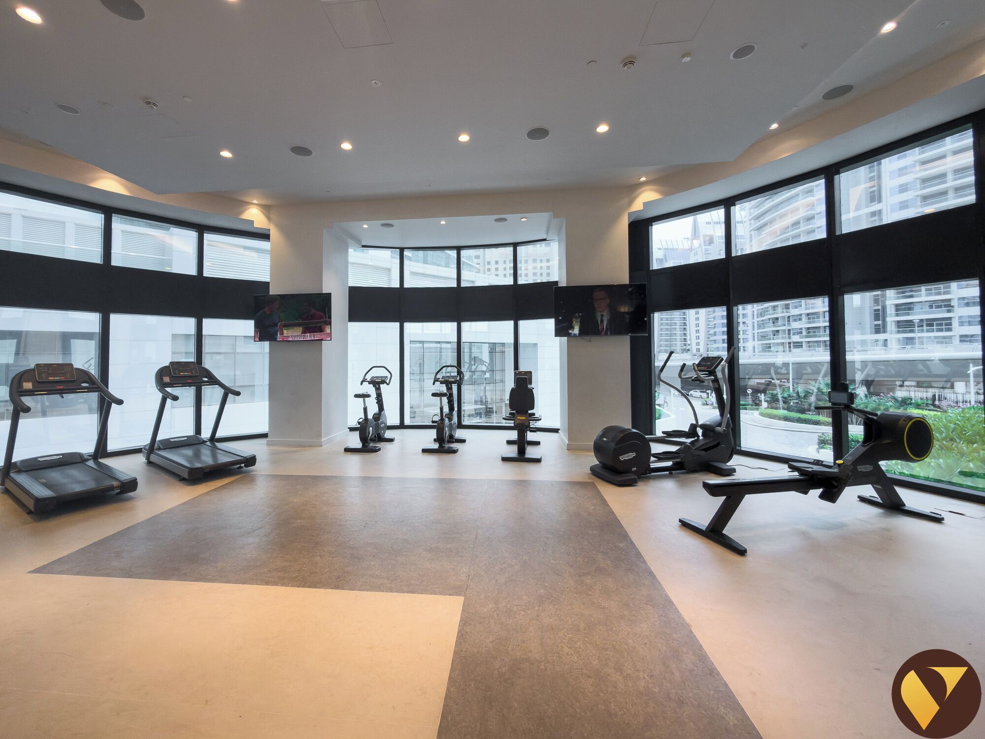 Une salle de sport moderne équipée de tapis de course, de vélos d’appartement et d’appareils de musculation, de grandes fenêtres offrant une vue sur la ville et d’un aménagement avec un sol plat.