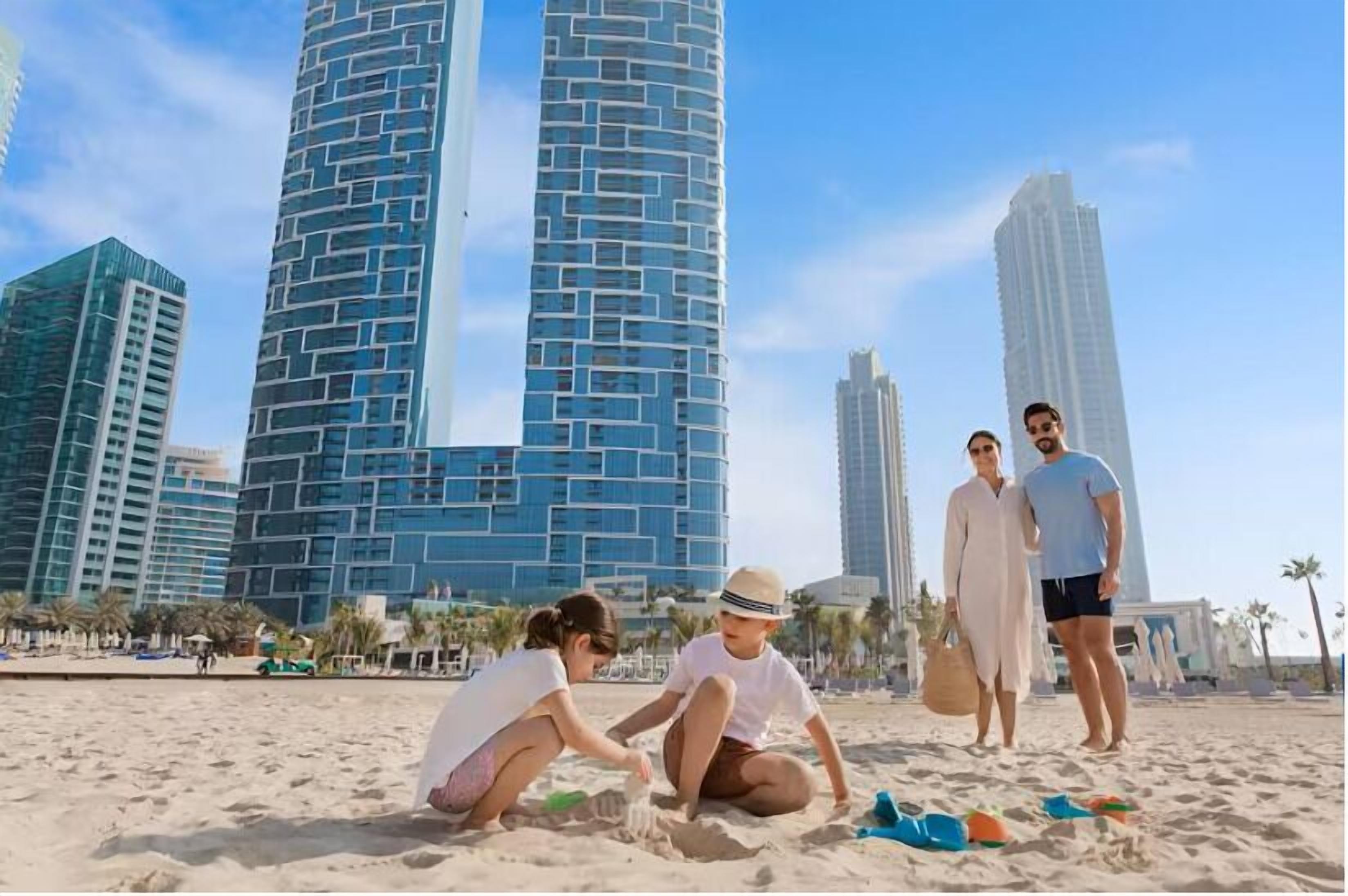 A beach with people, palm trees, and modern buildings in the background.