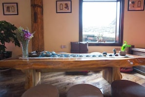 Une table en bois ornée de pierres et d’un vase de fleurs, une fenêtre donnant sur un paysage, et un canapé marron dans une pièce au plancher en bois.