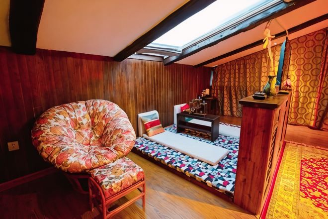 A cozy room with a wooden ceiling, a large floral chair, a small table, and a patterned rug.