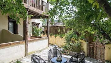 Eine Terrasse im Freien mit Korbmöbeln und einem kleinen Tisch, umgeben von Grünpflanzen und einem Gebäude mit einem Balkon.