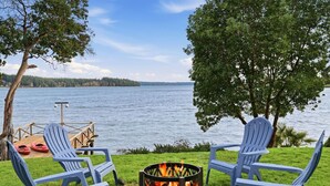 Vista al lago con dos sillas Adirondack, un pozo de fuego y un muelle con kayaks.