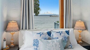 Habitación de hotel con cama, dos lámparas y vistas a una montaña y al agua a través de una ventana.