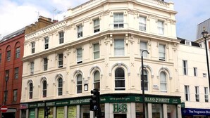 Um edifício de esquina com várias fachadas, incluindo a Hollands & Barrett, uma farmácia e uma loja de perfumes.