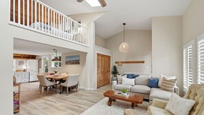 A spacious living area with a dining table, sofa, and a high ceiling with a skylight. - The Tide Runner Cottage | Short Walk to Beach + Tandem Kayak (Indianola)