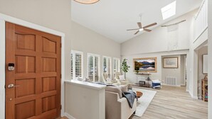 A spacious living room with a large sofa, a television, and a ceiling fan. - The Tide Runner Cottage | Short Walk to Beach + Tandem Kayak (Indianola)