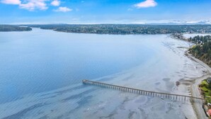 A coastal view with a pier extending into calm waters, surrounded by a forested landscape. - The Tide Runner Cottage | Short Walk to Beach + Tandem Kayak (Indianola)