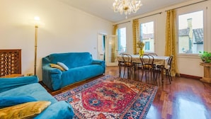 Een woonkamer met een blauwe bank, een eettafel met stoelen, een kroonluchter en een kamerbreed tapijt met een patroon.