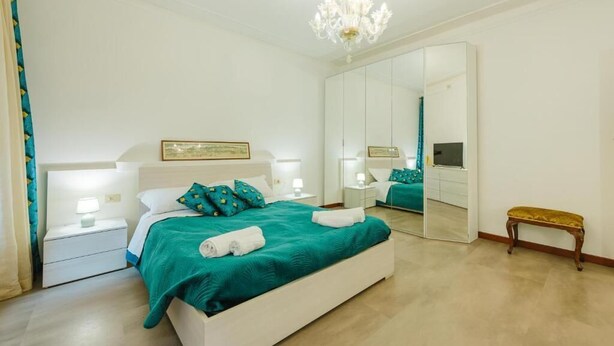 Een slaapkamer met een groot bed, nachtkastjes met lampen, een bankje, een spiegel en een kroonluchter.