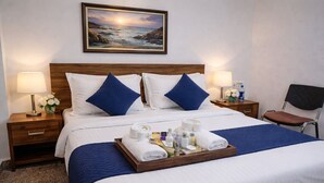 Habitación de hotel con cama, dos mesitas de noche, una silla, bandeja con toallas y artículos de aseo, y un cuadro en la pared.