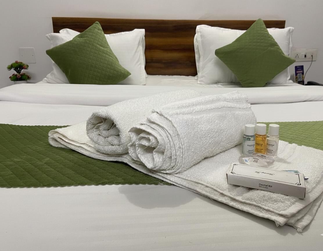 A neatly made bed with white bedding and green pillows, a rolled white towel, and a dental kit on the bed.