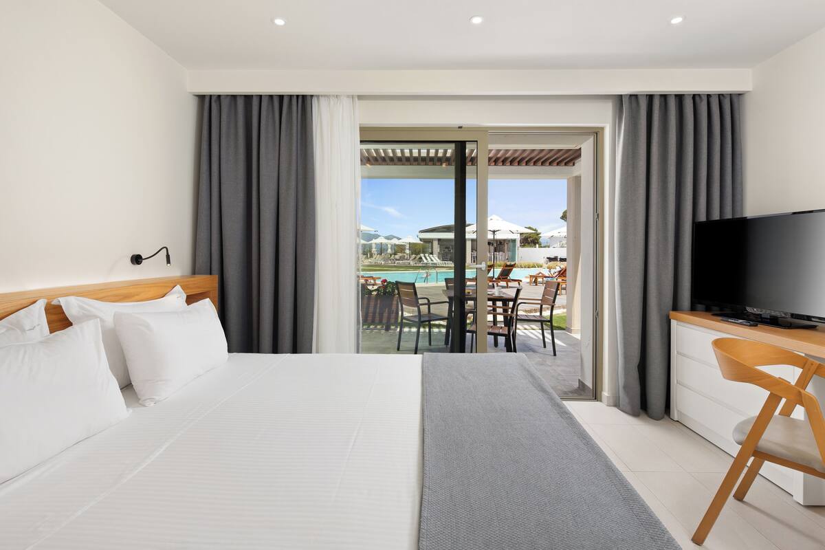 A hotel room with a bed, a TV, a desk, and a view of a pool and outdoor seating.