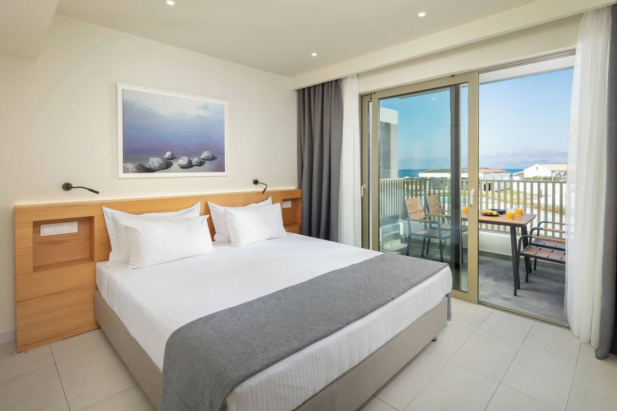 A hotel room with a bed, a painting, a balcony with a table and chairs, and a view of the sea.