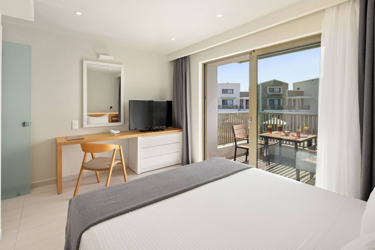 A hotel room with a bed, a desk with a chair, a television, a balcony with a table and chairs, and a view of buildings.