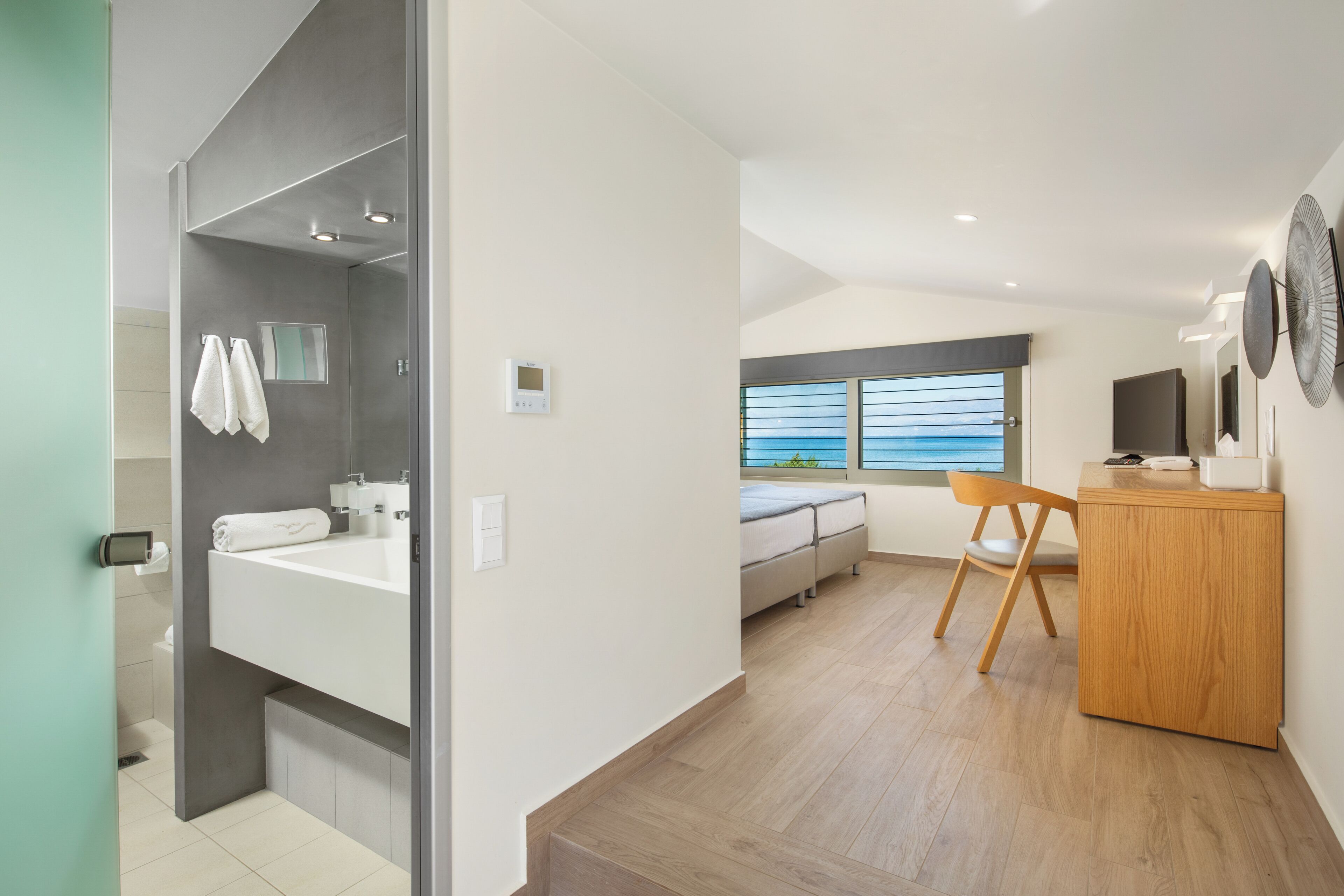 A modern hotel room with a large bathroom, a bed, a wooden desk, and a view of the sea.