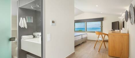 A modern hotel room with a large bathroom, a bed, a wooden desk, and a view of the sea.