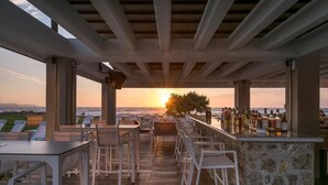 A covered outdoor bar area with tables and chairs, a stone wall, and a view of the sunset over the ocean. - Thalassa Suites Corfu (Corfu)