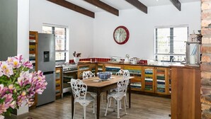 A kitchen with a wooden dining table, white chairs, a refrigerator, and a clock on the wall. - Narnia Luxury Self Catering Guest House (Knysna)