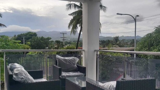 A covered outdoor seating area with wicker furniture, a glass railing, and a view of mountains and greenery. - Whitehaven Apartment in Lautoka, Fiji (Lautoka)