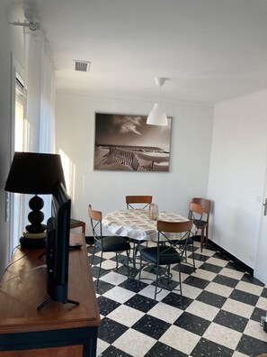 Uma sala de jantar com chão xadrez, uma mesa com cadeiras, um quadro na parede e uma luminária em uma escrivaninha.