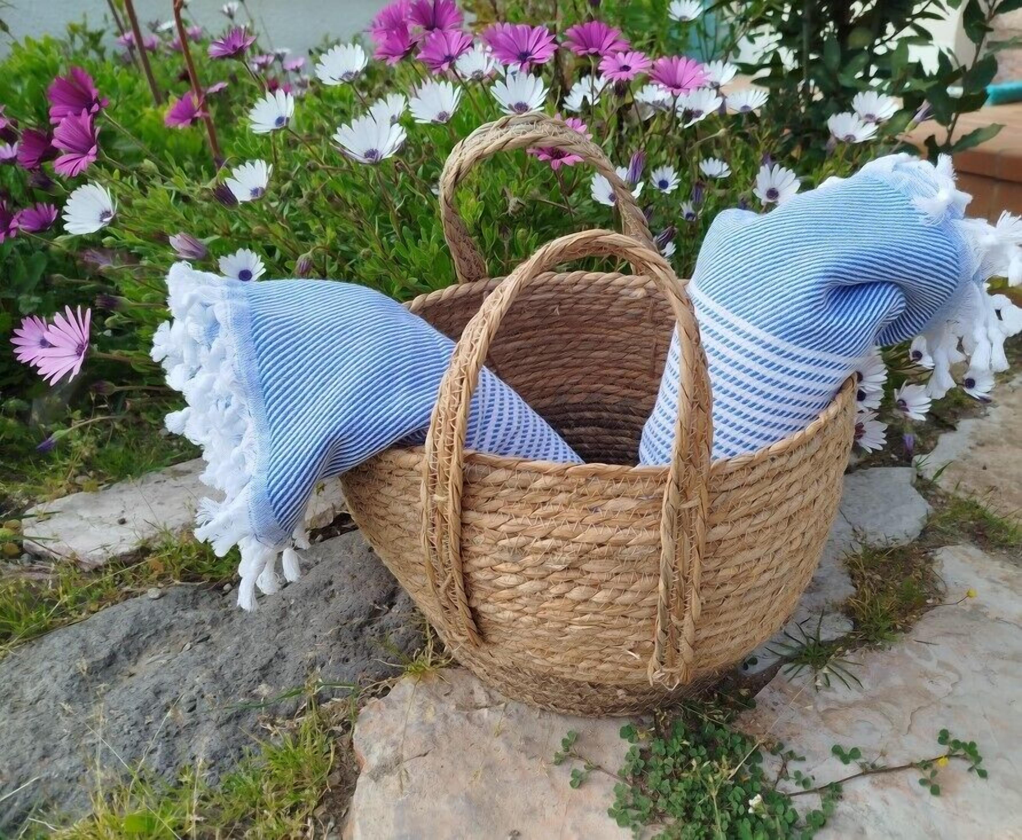 Ein geflochtener Korb mit Tragegriffen, gefüllt mit blau-weiß gestreiften Textilien, steht auf einem Stein mit Blumen im Hintergrund.